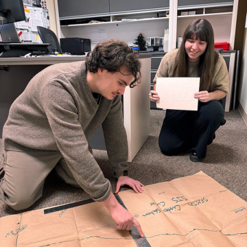 Engineering team members work on the floor using a full‑scale kraft paper template marked with field measurements for the curved expansion joint. One person references a metal ruler along the layout while another holds project notes.